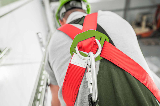 Travailleur vu de dos portant un harnais de sécurité rouge et vert correctement attaché à un point d’ancrage, dans le cadre d’une formation port du harnais