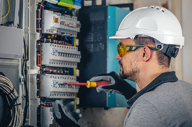 Électricien en formation BT intervenant sur un tableau basse tension avec casque, lunettes de protection et tournevis isolé