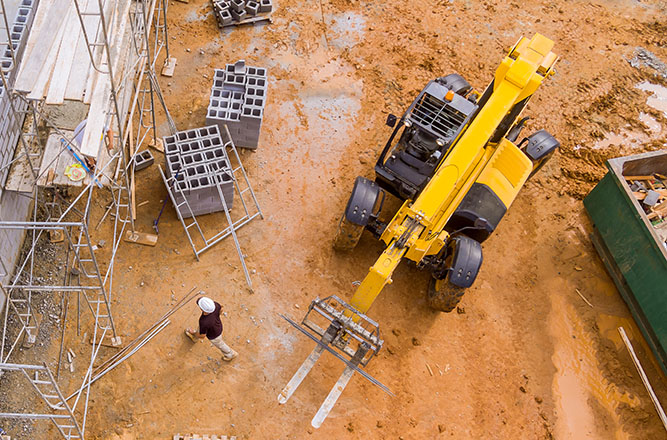 Chariot télescopique manœuvré sur un chantier en terre battue avec un opérateur au sol supervisant le levage de matériaux